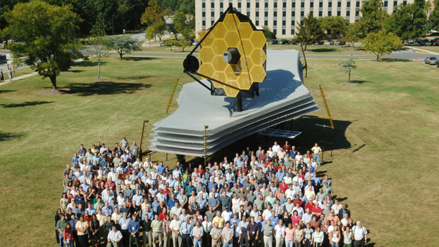 A crowd stands on a lawn in front of machinery adorned with solar panels.