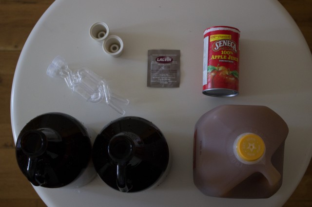 The materials of cider-making, clockwise from top left: two airlocks, two rubber bungs for stopping the fermentation vessels, a packet of yeast, apple juice concentrate, apple cider, two growlers.