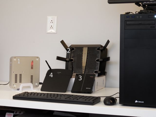 Clockwise from the far left: 1. the Ubuntu-powered Homebrew Special, 2. the Netgear Nighthawk X6, 3. the Buffalo WHR-G300N-v2, and 4. the Linksys N600 EA-2750. Far right, looming over them all: Monolith, one of the two servers used for testing.