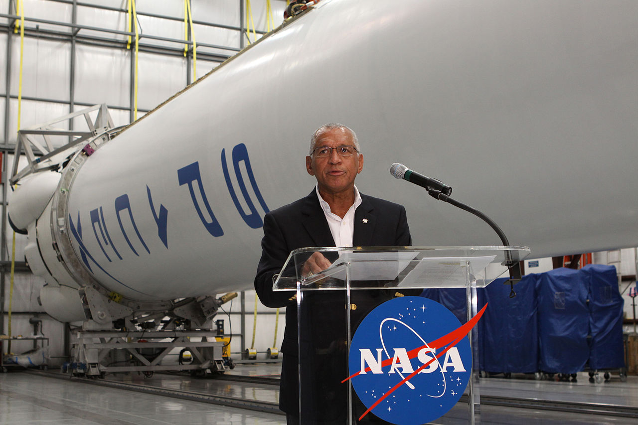 Man with glasses speaking behind a NASA podium in front of a space ship.