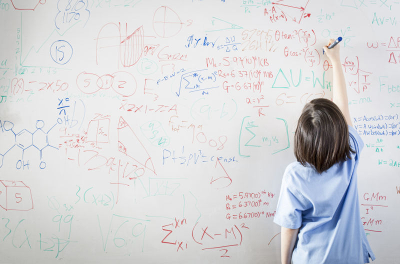 A child writes on a whiteboard cluttered with equations.