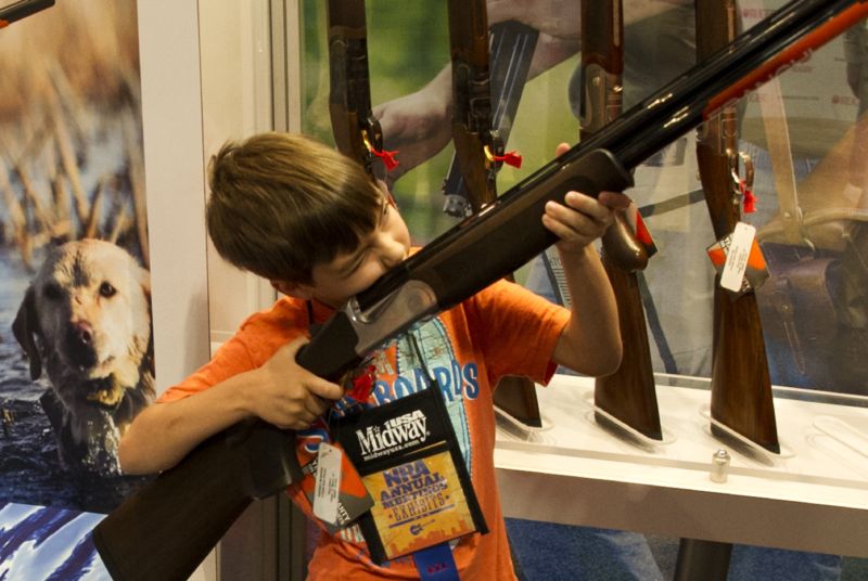 A boy under his parents’ supervision aims a shotgun.