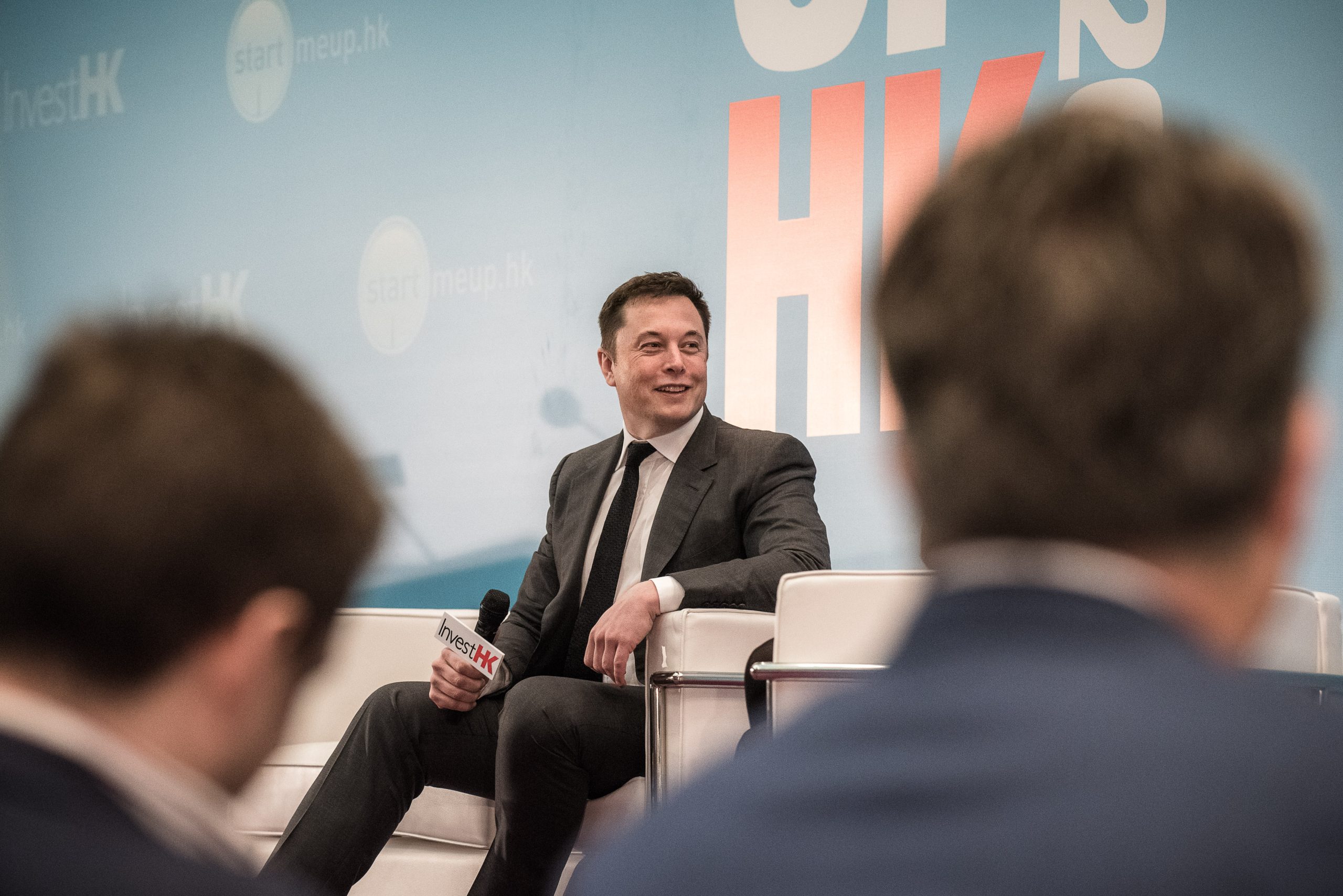 A man in a suit sits with a microphone on a stage.