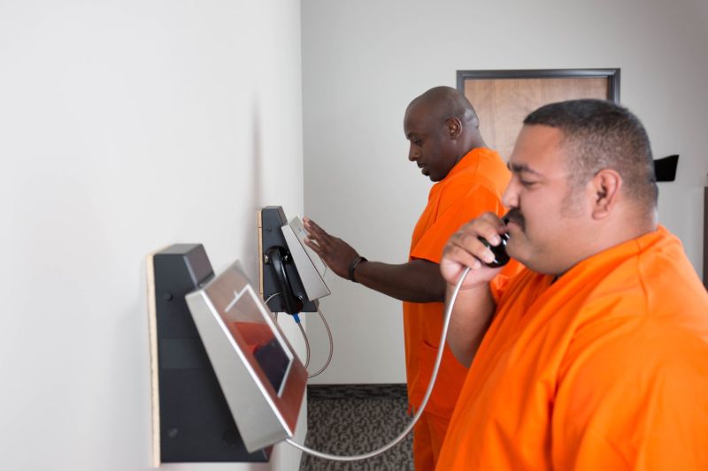 Prison inmates in orange jumpsuits use video-visitation kiosks.