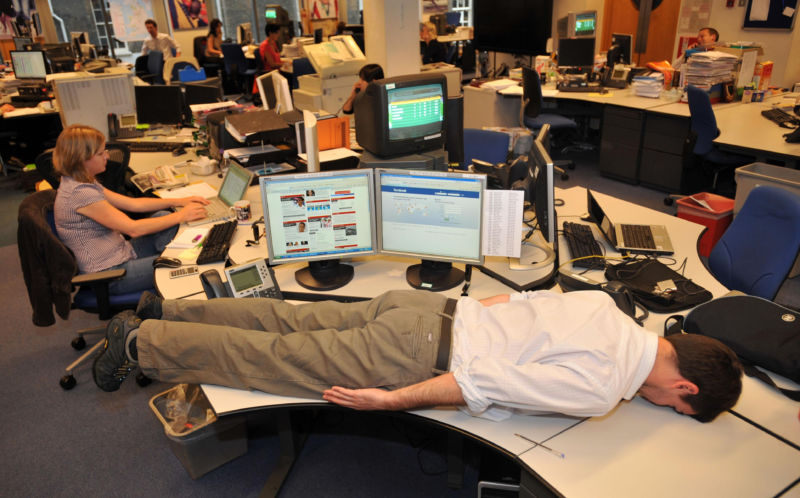 A man lies face down on a desk in an open office