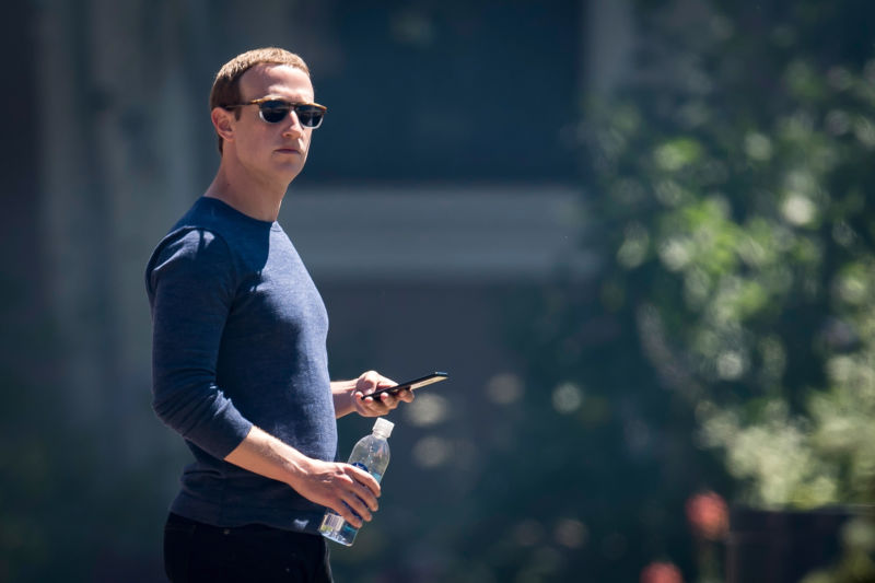 A man in glasses is holding a cell phone and a bottle of water while standing outside.