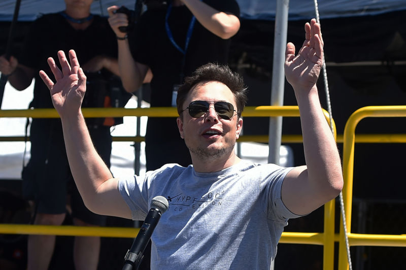 Man in T-shirt and sunglasses waves at crowd before speaking into microphone.