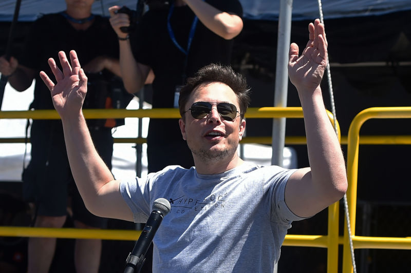 A man in a t-shirt and sunglbades greets the crowd before speaking into the microphone.