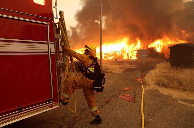 A firefighter pulls a hose away from a horse barn as a fire rages in the background.