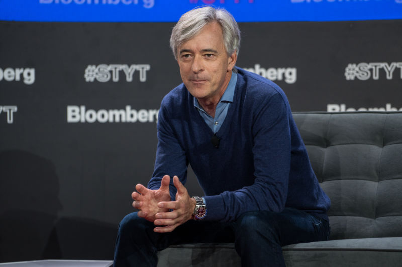 A middle aged man sits on a stage during a presentation.