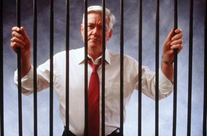 A man with white hair, wearing a button-down shirt and tie, standing behind the bars of a jail cell.