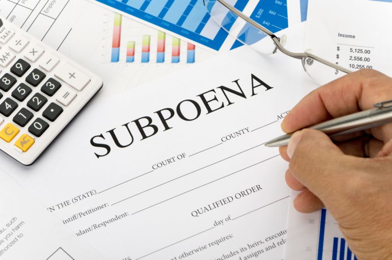 A person's hand holding a pen and filling out a subpoena form.