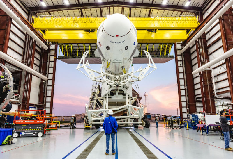 A man in a helmet stands in front of a giant rocket in an even bigger hanger.