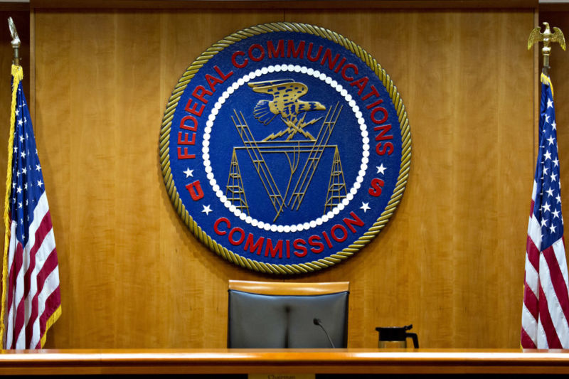The Federal Communications Commission meeting room, with an empty chair in front of the FCC seal and two United States flags.