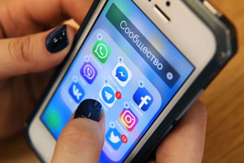 A woman holds a smartphone with icons for social networking apps Facebook, Instagram, Twitter and others. The app icons are in a folder whose title is the Russian word for 