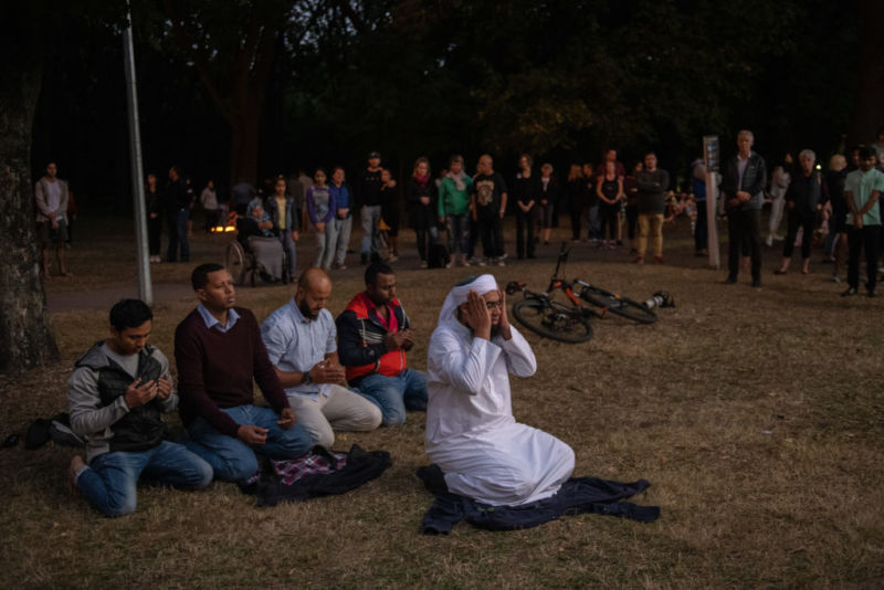 People look on as men pray in a park near Al Noor mosque after a terrorist killed 50 people.