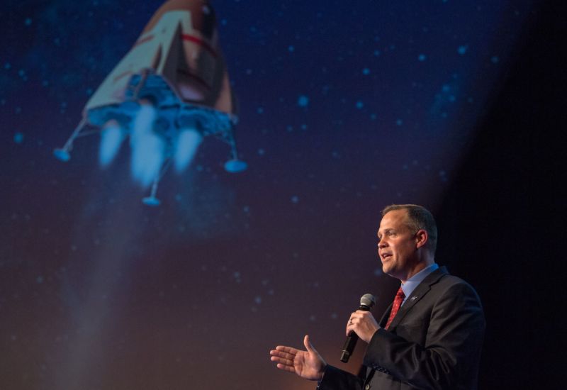 A man in a suit speaks into a microphone in front of a large image of a spacecraft.