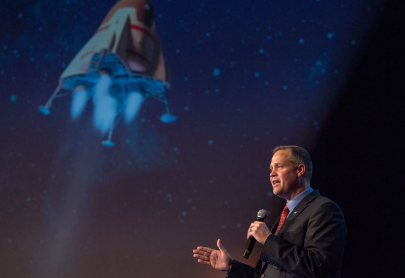 A man in a suit speaks in a microphone in front of a large image of a spaceship.