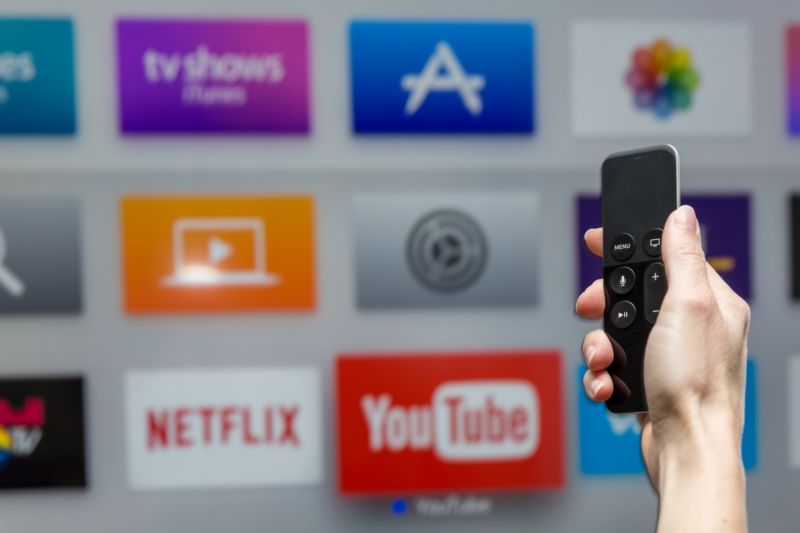 A person's hand holding an Apple TV remote in front of a TV screen displaying the app icons for YouTube and other services.