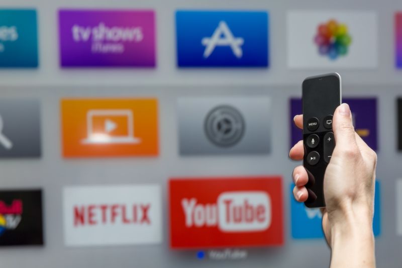 The hand of a person holding an Apple TV remote control in front of a TV screen displaying application icons for YouTube and other services.