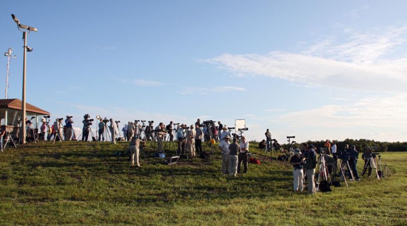 People with cameras and tripods crowd a grassy field, all facing the same direction.