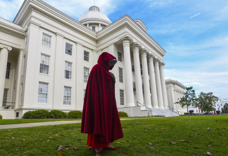 A woman in a red hooded cloak stands in front of a Neo-Federalist building.