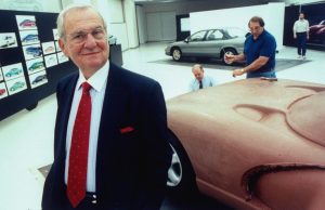 Lee Iacocca posing in front of full-sized clay model of the proposed Viper sports car being worked on by staff technicians in the Advanced International Design studio at the Chrysler Tech Center.