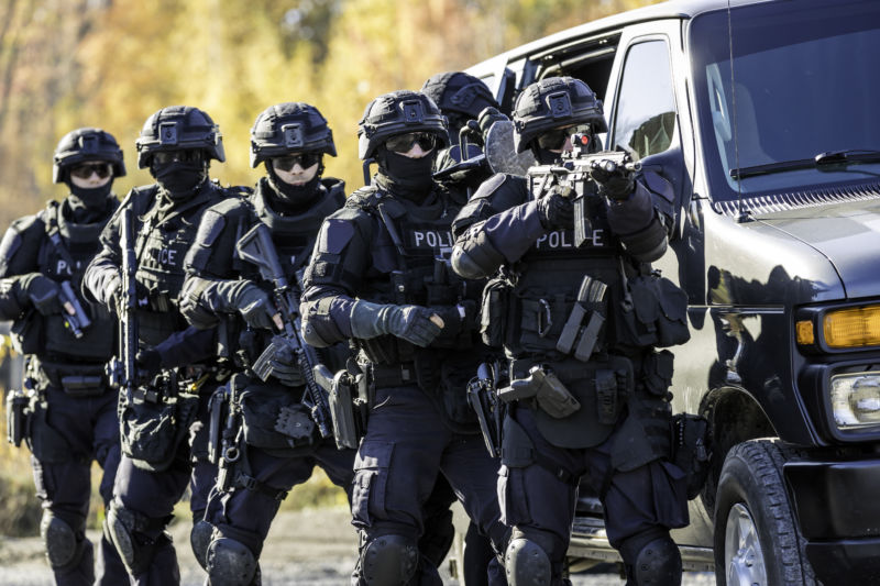 Heavily armed men in body armor stand ready behind a van.