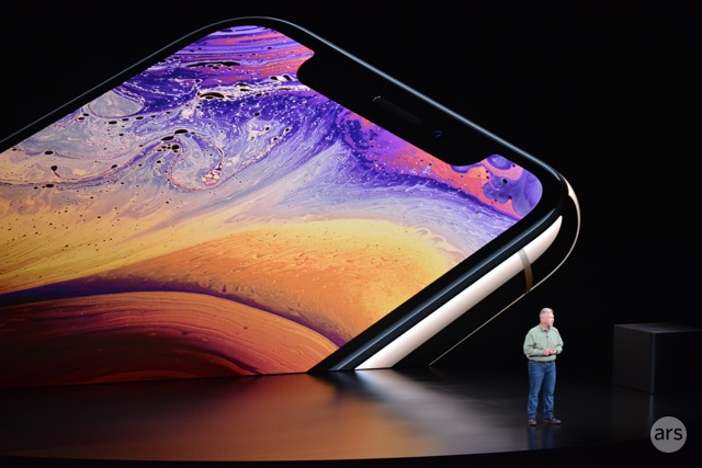 A man on a stage is dwarfed by a gigantic video display of a smartphone.