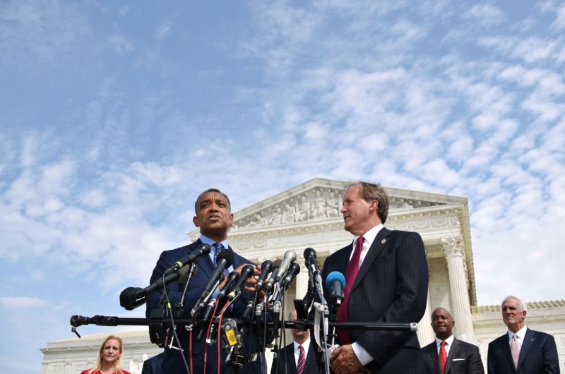 Two men stand in front of an array of microphones before a Federal-style federal building.