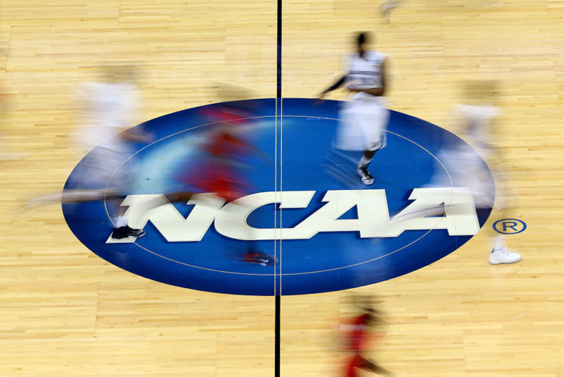 College basketball players running on top of the NCAA logo on a basketball court.
