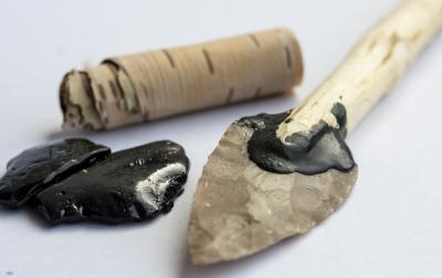 Color photo of a roll of birch bark, a puddle of tar, and a spear with birch tar securing the point.