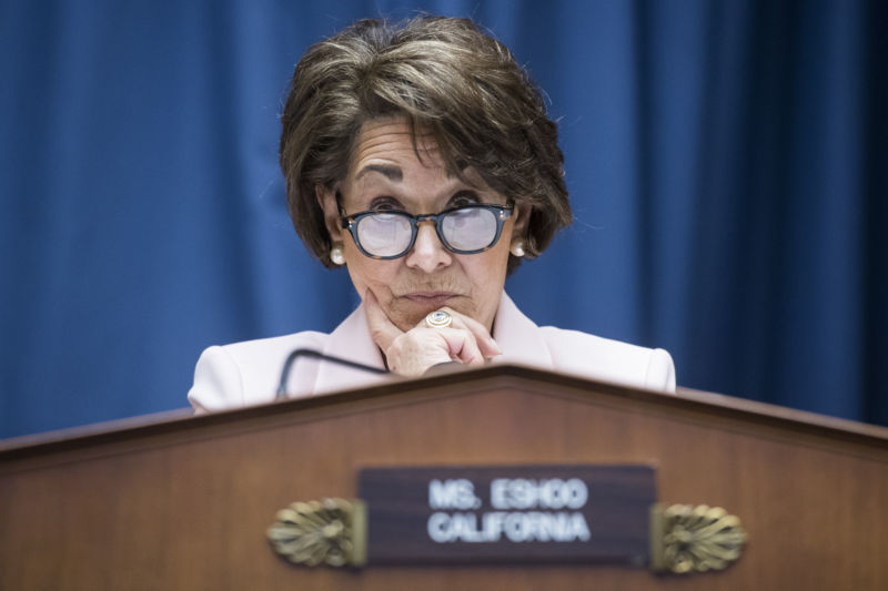 A serious woman in a business suit listens at a podium.