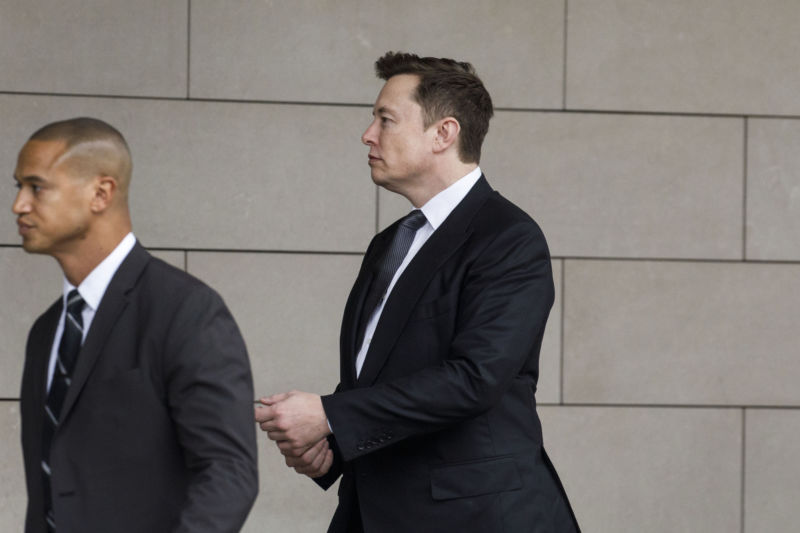 Men in suits walk past a brick wall.