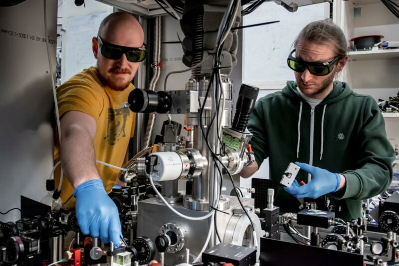 Image of two scientists wearing gloves and sunglasses in amidst lab equipment.