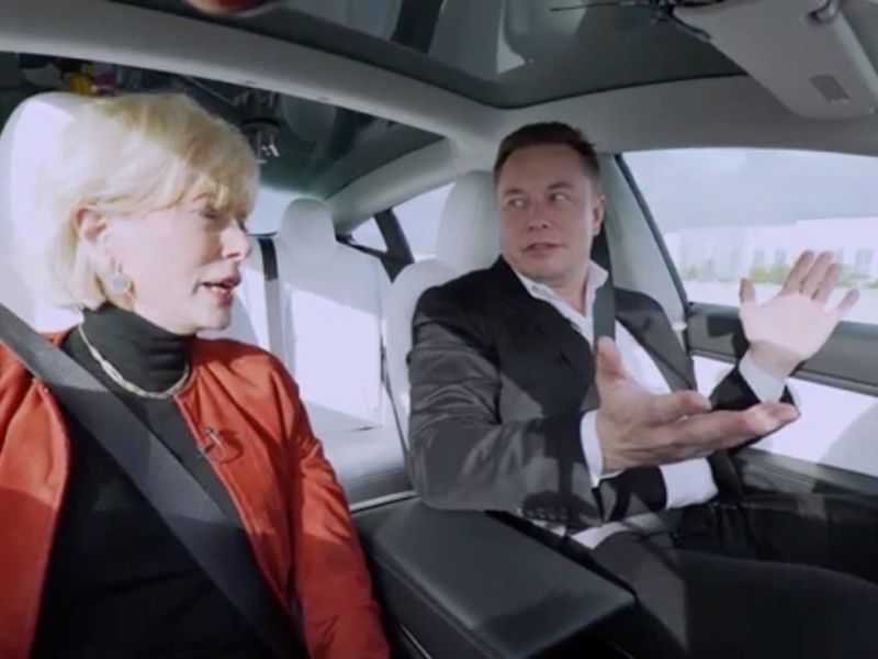 Elon Musk and Barbara Walters in a Tesla. Musk has his hands off the steering wheel as the car is driving.