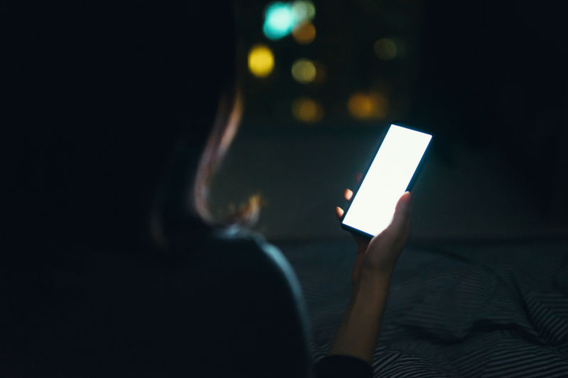 Young woman online shopping on smartphone while lying in bed in darkness at night