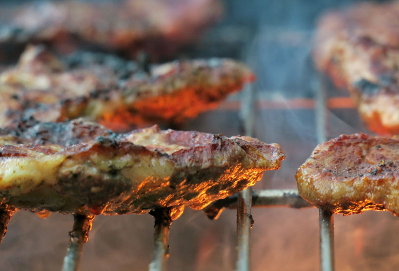 A close-up of charred meat cooking on a grill with flames below and rising smoke