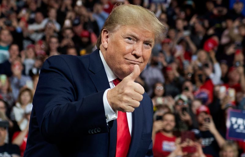 A picture of President Trump in a blue suit, with a red tie, looking at the camera while giving a thumbs-up. A crowd of supporters are seen, out of focus, behind him.