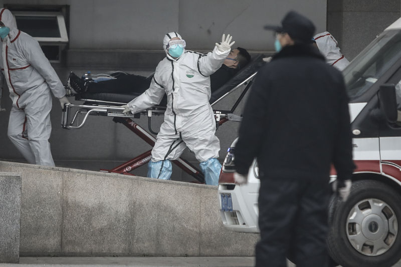 A person in a white protective suit, a blue face mask and glasses helps to send a patient on a gurney to a hospital.  His hand is outstretched as if signaling someone not to come near.