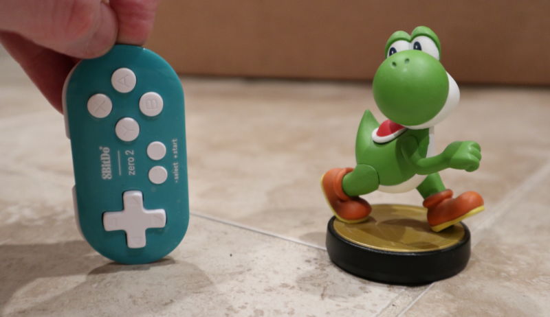 A hand holds a tiny video game controller next to a dinosaur figurine.