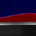 A sand dune constructed from red glass beads. Some of the beads are being blown off by water flow.