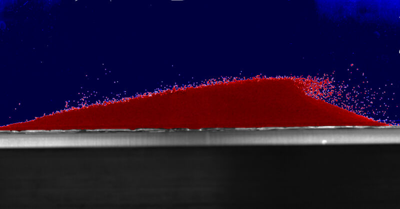 A sand dune constructed from red glass beads. Some of the beads are being blown off by water flow.