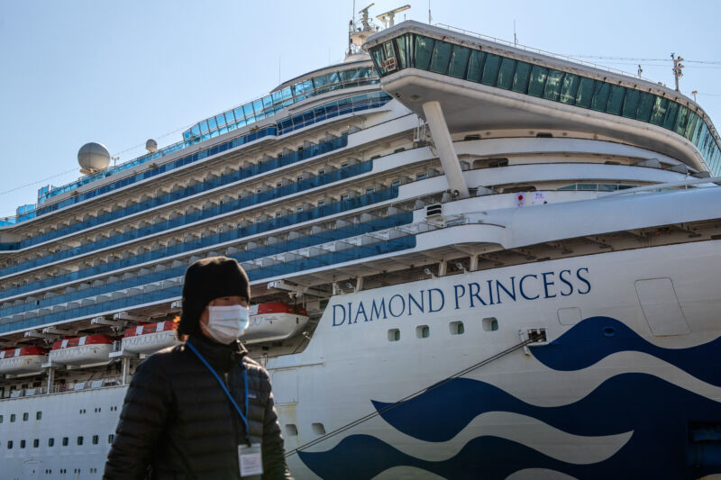 A person wearing a face make walks along a port on a sunny day with the Princess Diamond cruise ship docked in the background.