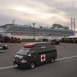 An ambulance sits in a parking lot in front of a docked cruise ship.