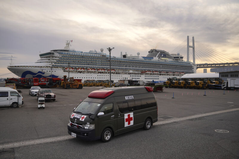 An ambulance sits in a parking lot in front of a docked cruise ship.