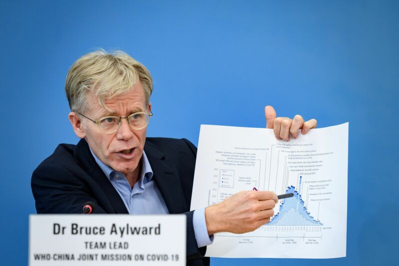 A harried man in an open-collar suit points at a graph on a sheet of paper.