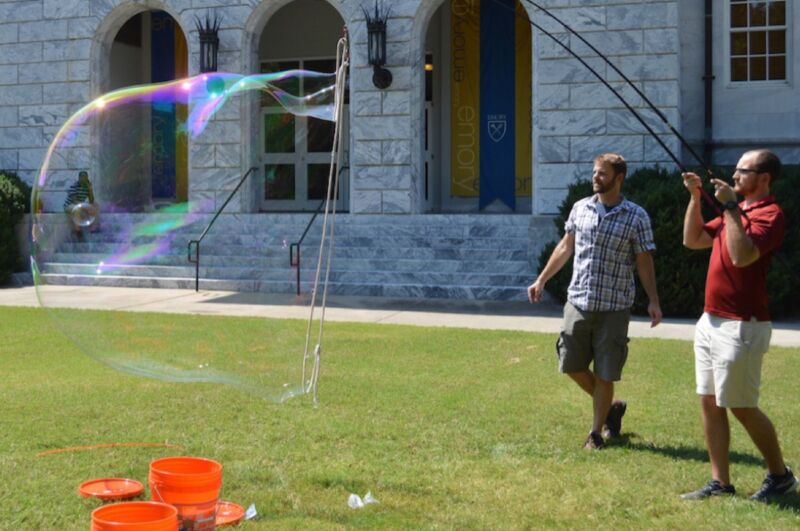 Two grown men blow giant bubbles on a lawn.