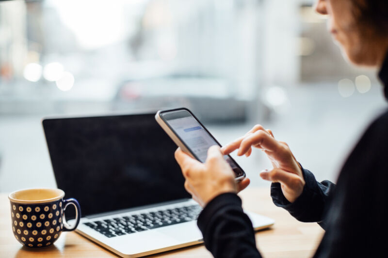 Stock photo of hands using smartphone with laptop in background.