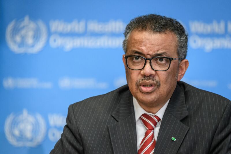 A serious man in a suit speaks in front of a blurry World Health Organization logo.